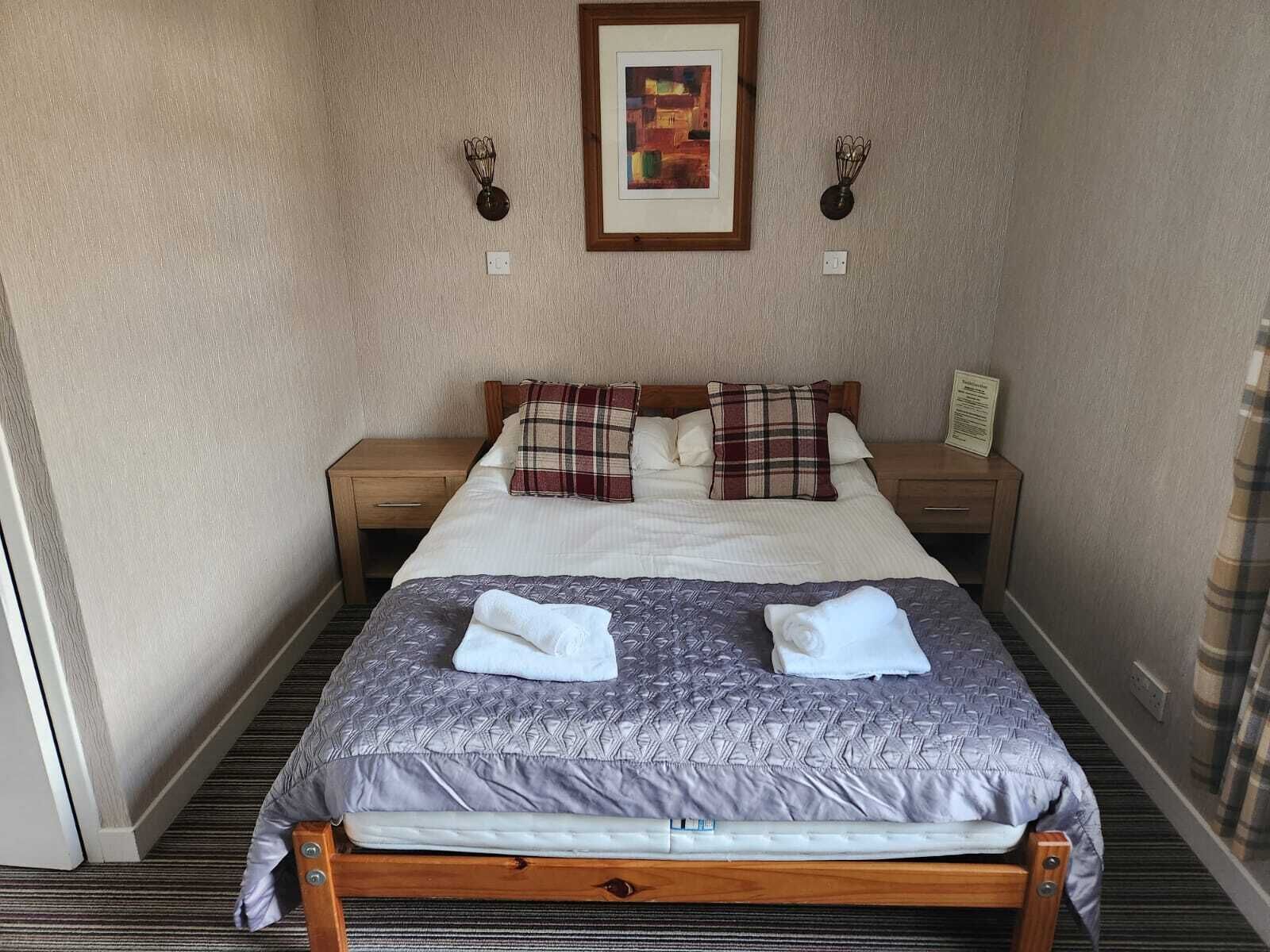 A neatly made bed with a plaid blanket and pillows, a framed picture on the wall, and a small table with a lamp beside the bed.