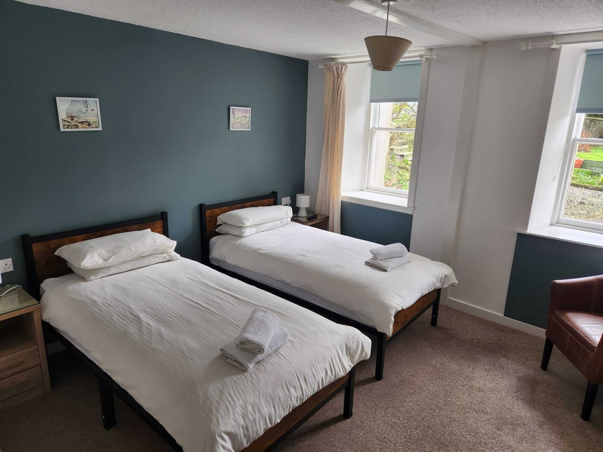 A room with two single beds, a wooden nightstand, a chair, and a window with curtains.