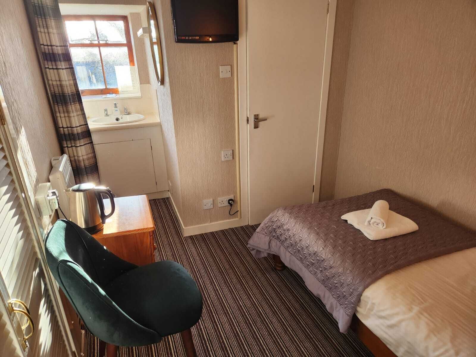 A hotel room with a bed, a chair, a desk with a kettle, a television, and a bathroom visible through an open door.