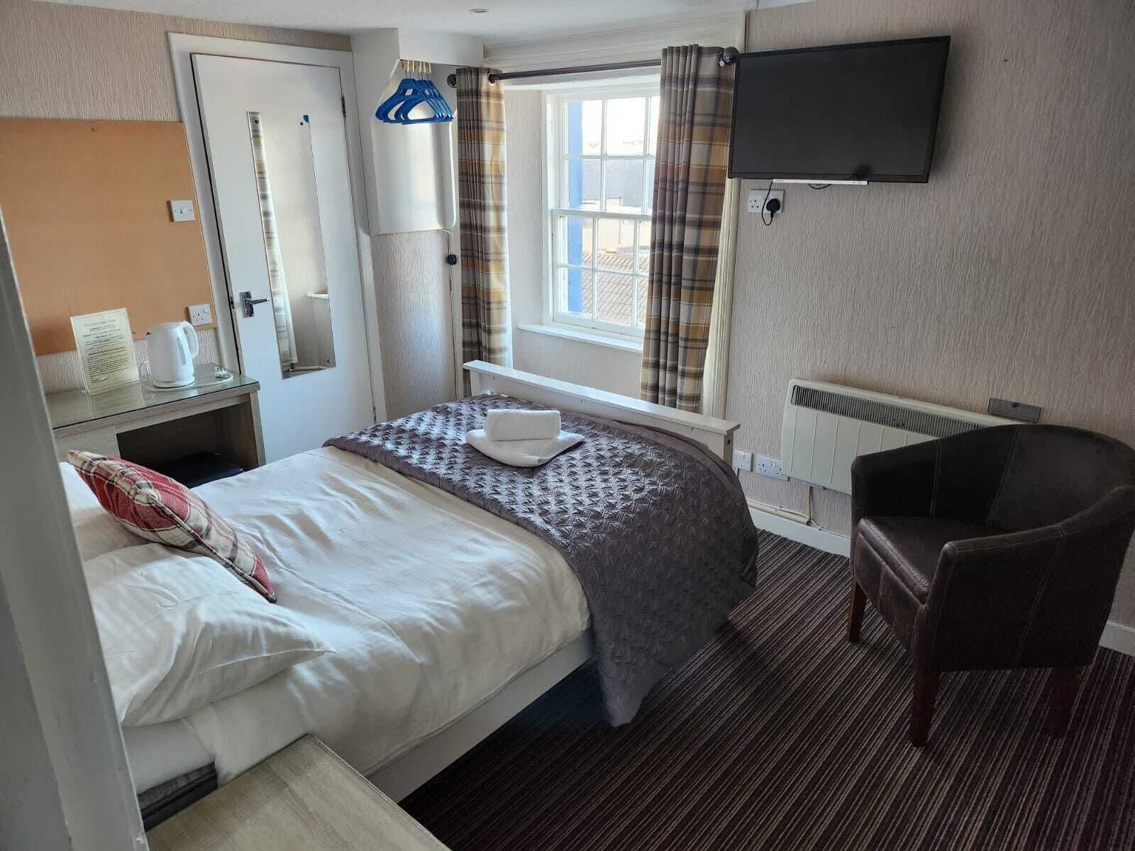 A hotel room with a bed, a chair, a TV, a window with curtains, a radiator, and a bedside table with a kettle.