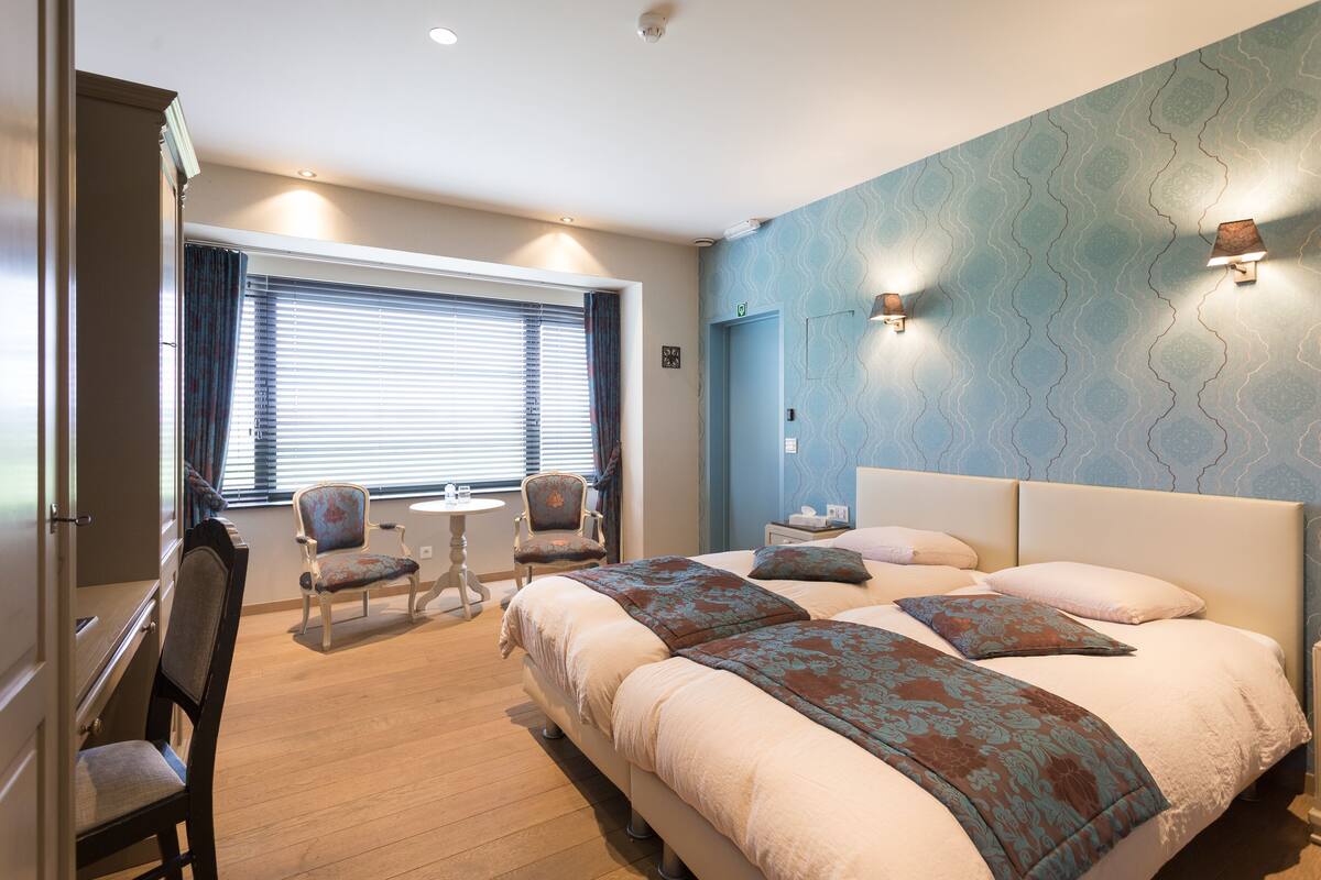 A hotel room with a large bed, two chairs, a small table, and a window with blinds.