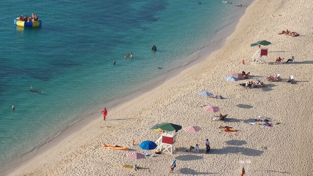Ein Strand mit klarem türkisblauem Wasser, einem Steg und mehreren Strandsonnenschirmen.