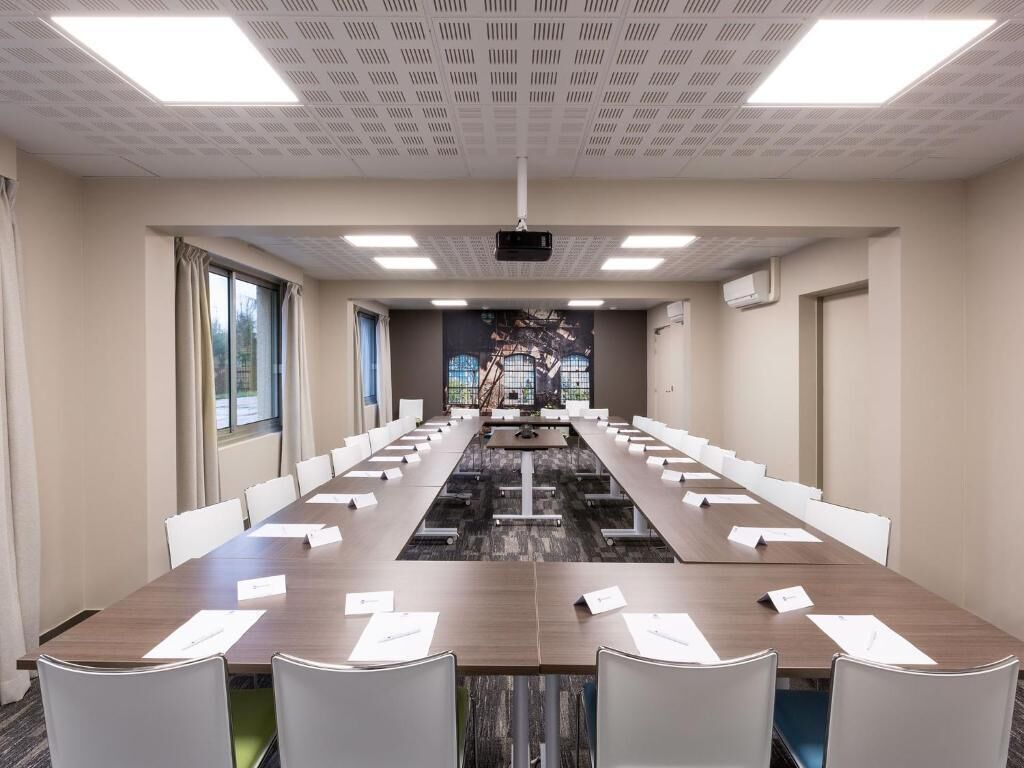 Ett konferensrum med ett långt bord, vita stolar, en projektor och ett stort fönster med gardiner.