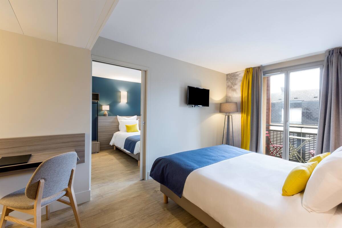 Une chambre d’hôtel avec un lit, un bureau, une chaise, une télévision et un balcon avec des rideaux.
