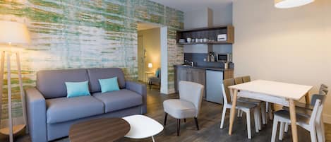 A modern living room with a sofa, a dining table, and chairs. There is a kitchen area with cabinets and a microwave.
