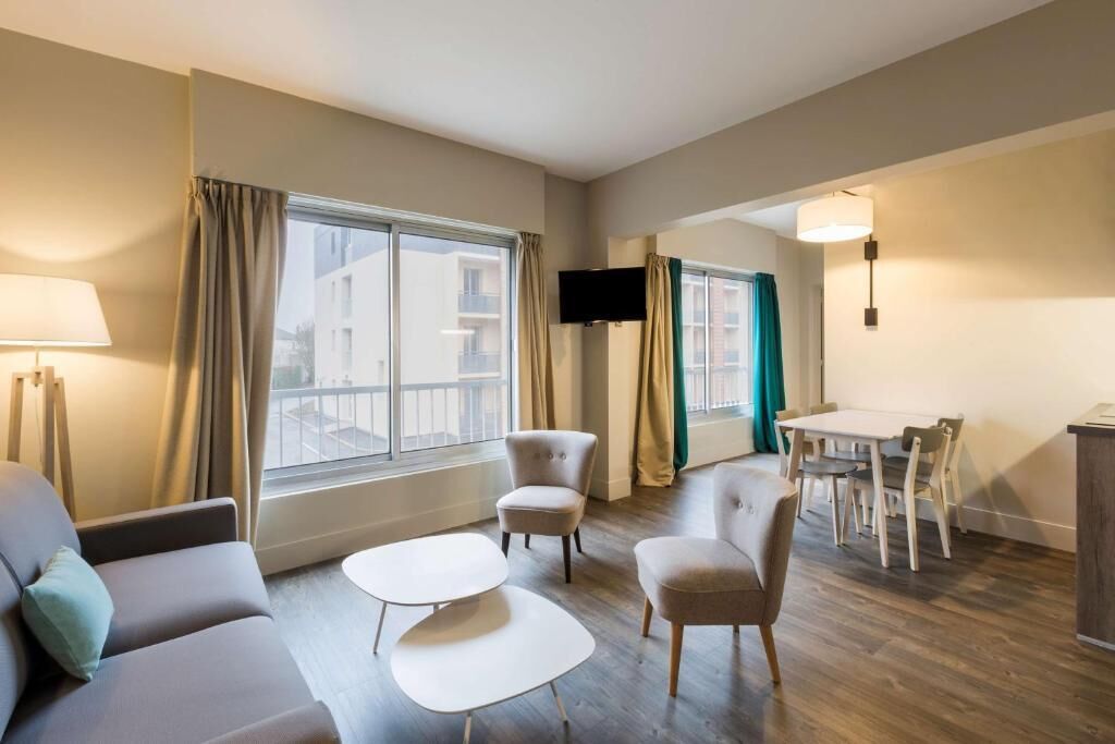 A modern hotel room with a sofa, two armchairs, a small table, a TV mounted on the wall, and a dining area with a table and chairs.