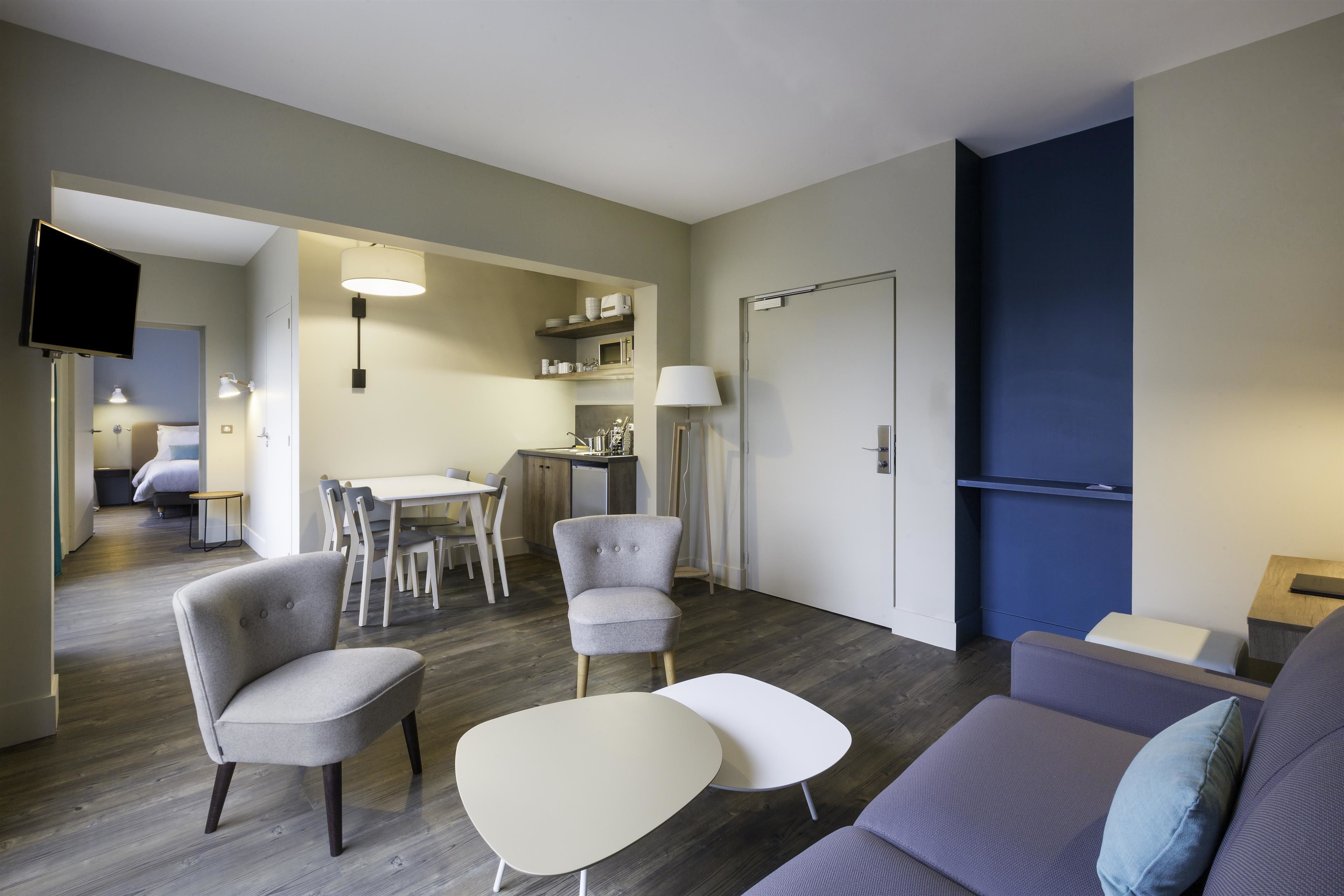 A modern living room with a sofa, two armchairs, and a coffee table. There is a dining area with a table and chairs, and a kitchen area with a microwave and shelves. A flat-screen TV is mounted on the wall.