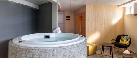 Un spa moderno con una bañera de hidromasaje con mosaicos, una pared de madera, una mesita pequeña y una silla con un cojín de colores.