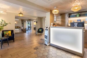 Un espace d’accueil d’hôtel au design moderne, comprenant un comptoir d’accueil, un coin salon avec un canapé et une petite table, ainsi qu’une plante décorative.