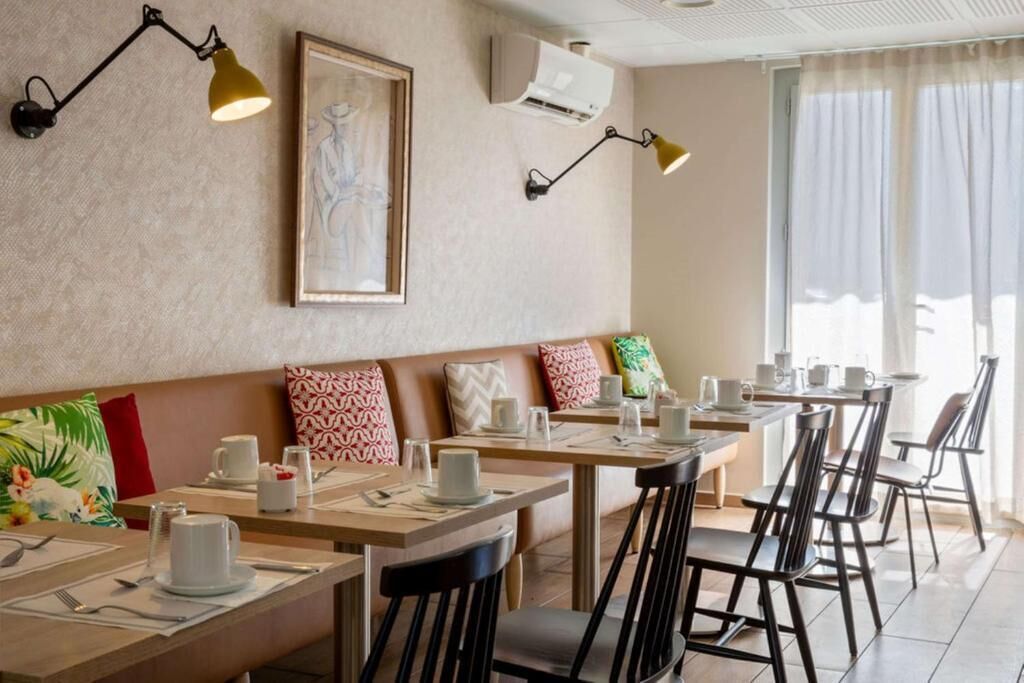 Ein Restaurant mit Holztischen und schwarzen Stühlen, eine Bank mit gemusterten Kissen sowie Wandleuchten.