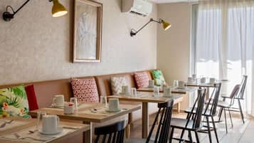 Ein Restaurant mit Holztischen und schwarzen Stühlen, eine Bank mit gemusterten Kissen sowie Wandleuchten.