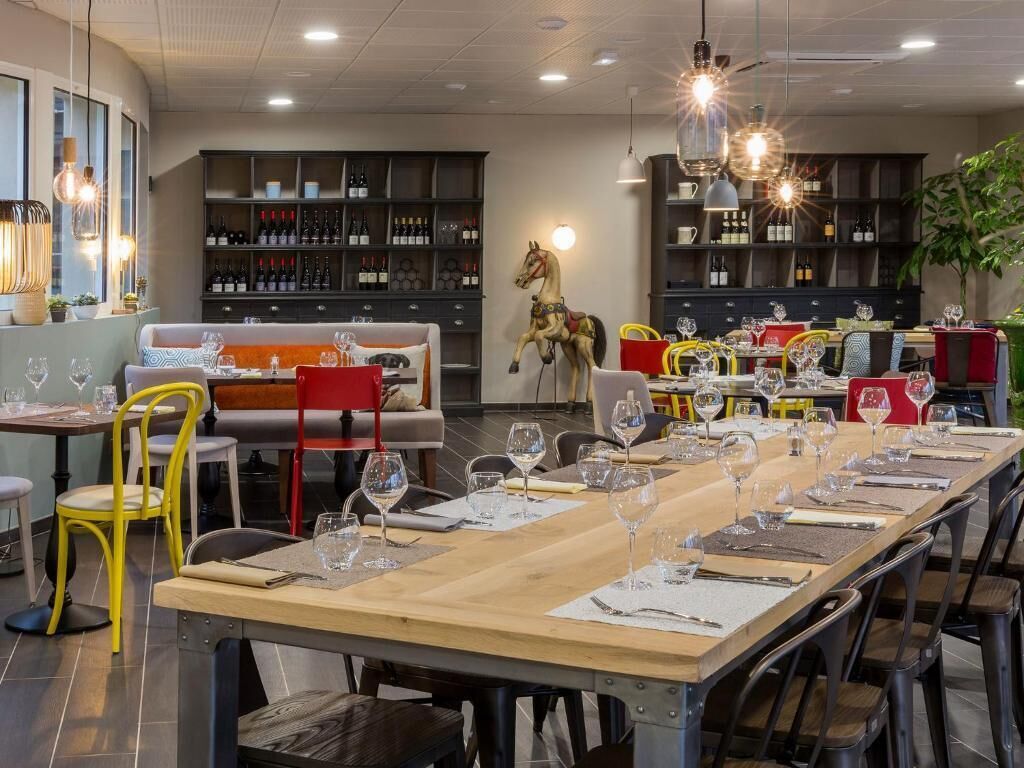 Un restaurant avec une longue table en bois, dressée pour le repas, entourée de chaises. On peut voir des verres à vin sur la table, et une sculpture de cheval est visible à l’arrière-plan.