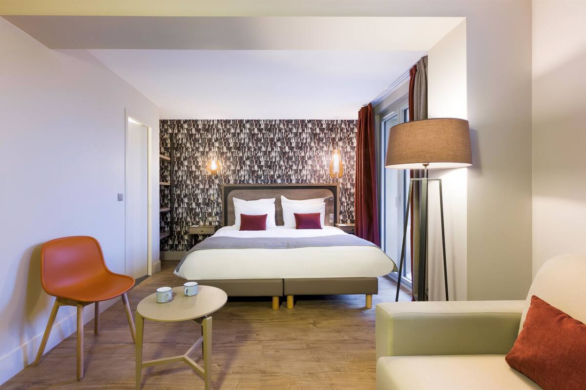 Une chambre d’hôtel moderne avec un grand lit, un bureau avec deux chaises, une petite table et une lampe sur pied.