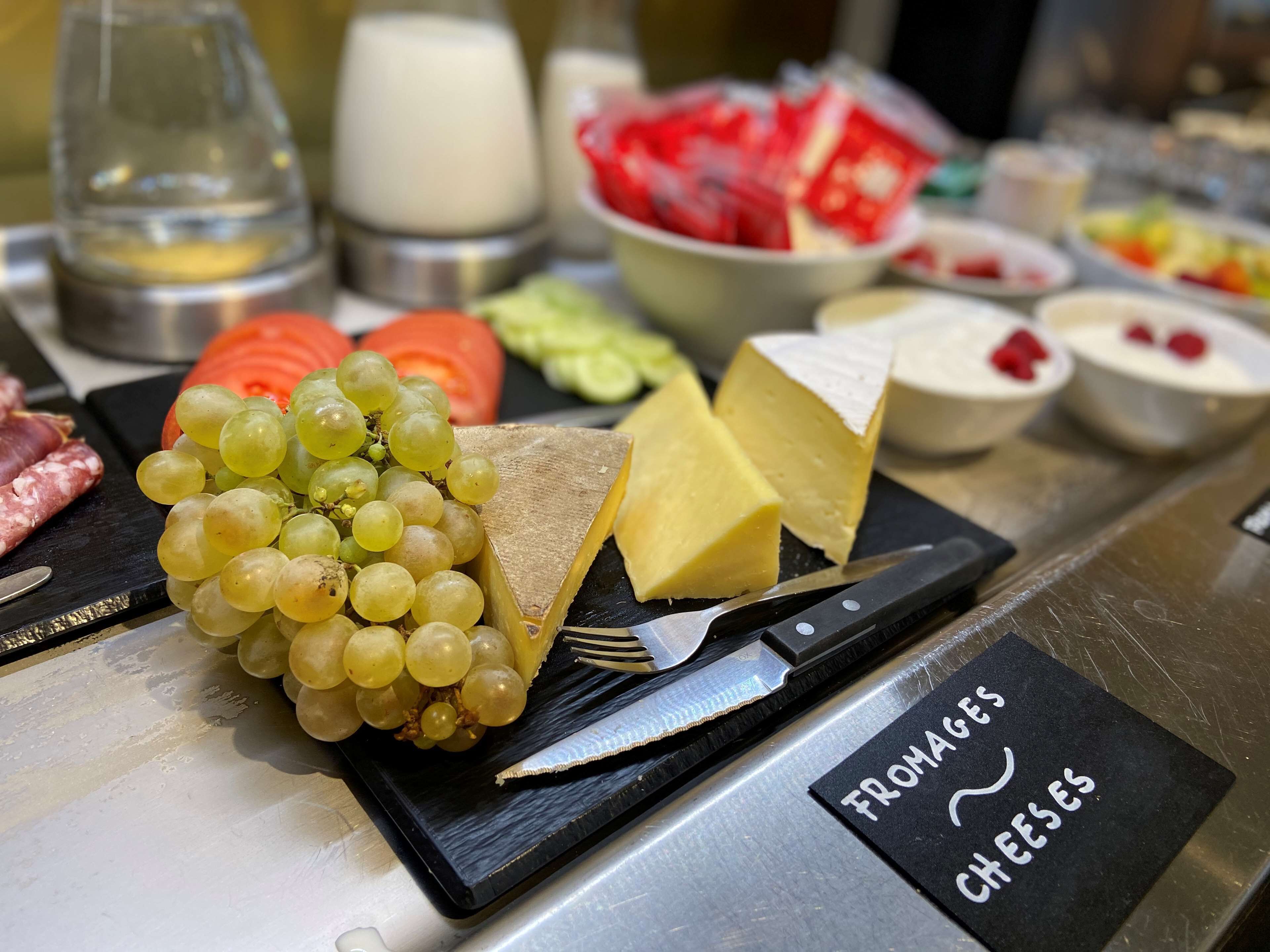 Una tabla de quesos y frutas con un cuchillo y un tenedor sobre una losa negra, con la etiqueta 'FROMAGES CHÈES'.
