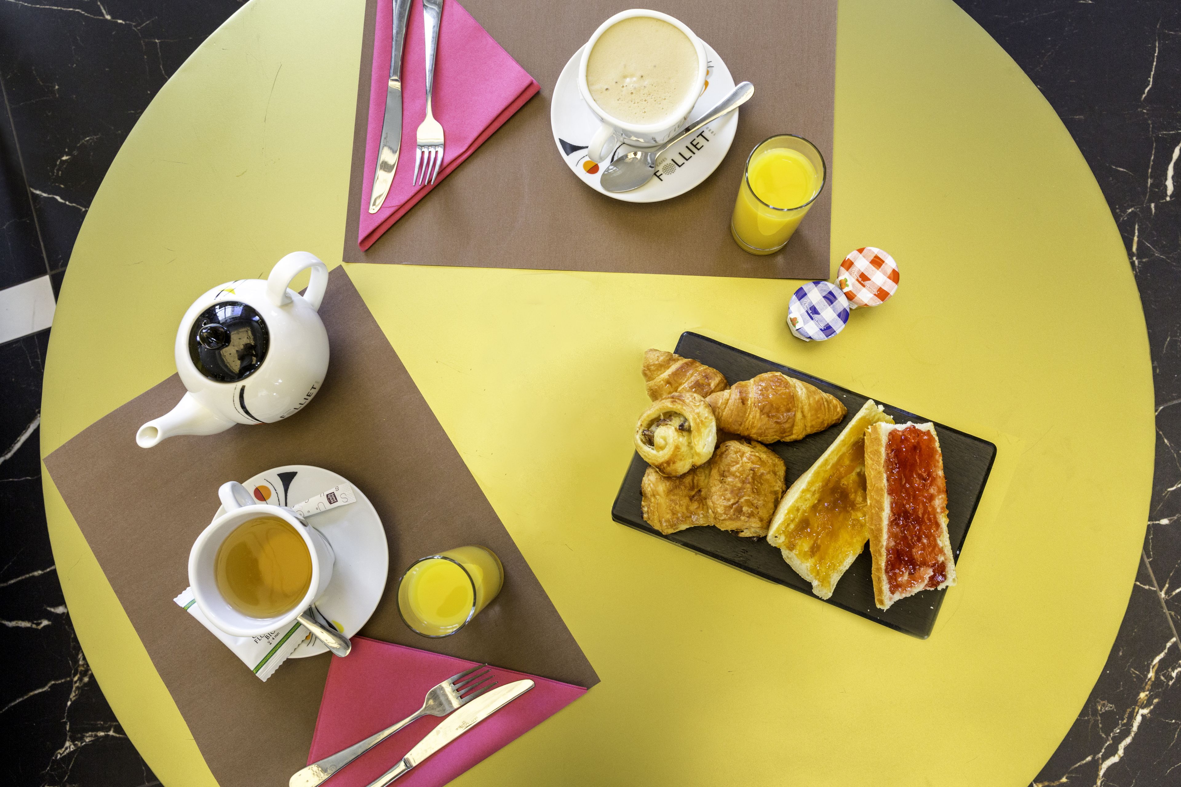 A breakfast setting with a teapot, cup, croissants, and juice on a table.