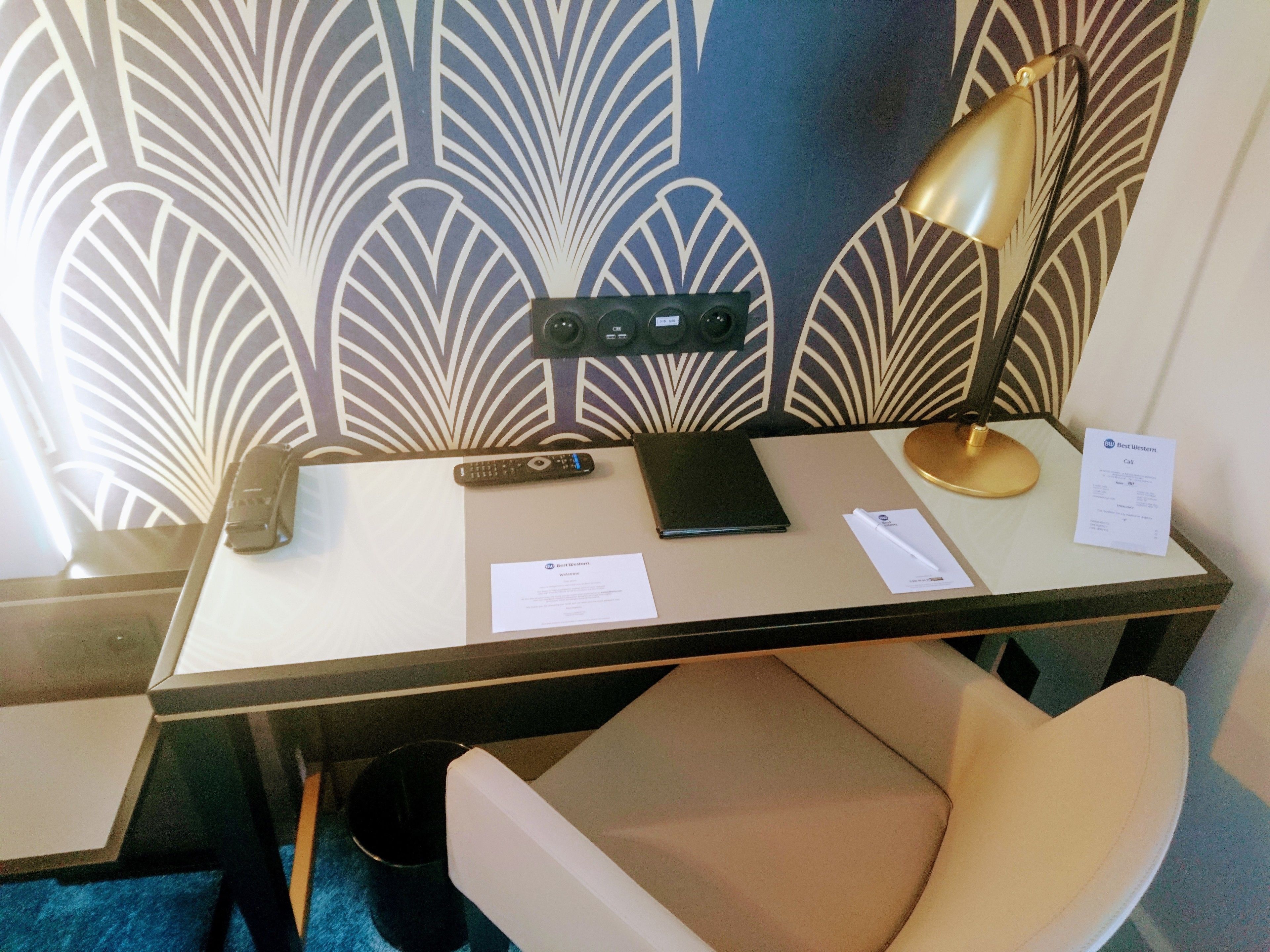 Une chambre d’hôtel équipée d’un bureau, d’une télécommande, d’une lampe, d’une chaise et d’un mur décoré d’un motif.
