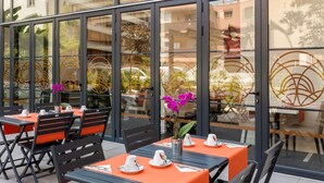 Un área de comedor al aire libre con mesas y sillas, ventanas amplias y vistas a edificios y árboles.