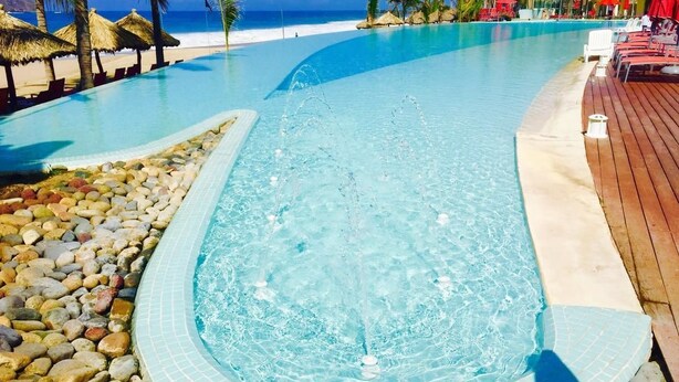 Una piscina azul cristalina con cubierta de madera y sillas de descanso, rodeada de palmeras y vista a la playa.