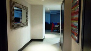 Un pasillo con un espejo moderno, un cuadro abstracto y colorido, y una vista parcial de una habitación con cama y sillón.