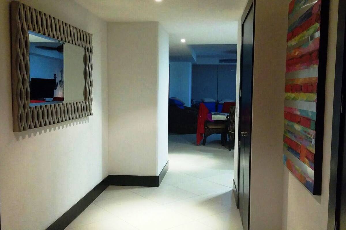 A hallway with a modern mirror, a colorful abstract painting, and a glimpse of a room with a bed and a chair.