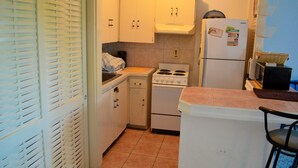 A compact kitchen with white cabinets, a stove, a refrigerator, and a small dining area with a chair. - Crystal Beach Studio (Montego Bay)