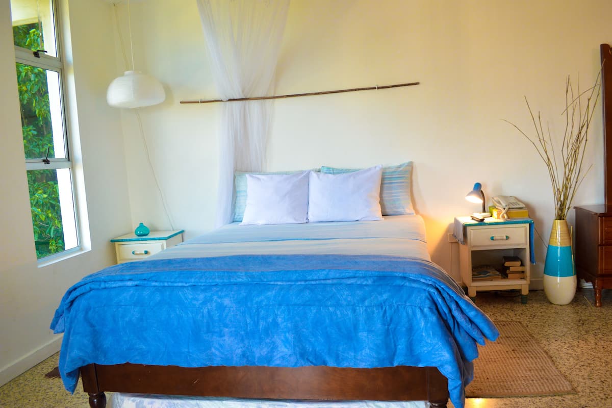 A bedroom with a bed, bedside tables, a lamp, a nightstand, a window, and a vase with plants.