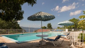 Una piscina con lettini e ombrelloni, circondata da un recinto bianco e alberi.