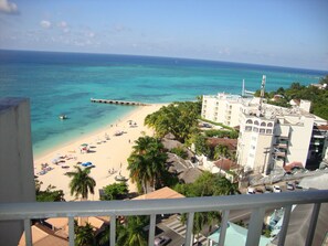Una spiaggia con acque cristalline turchesi, un molo che si protende verso il mare e un hotel con diversi piani e balconi affacciati sulla spiaggia.