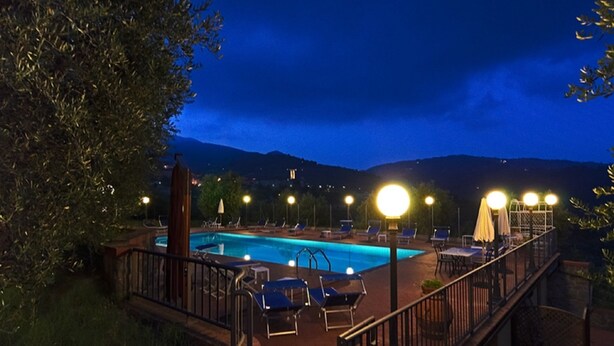 Vista nocturna de una piscina con sillas de descanso y mesas, rodeada por una terraza con barandillas y farolas.