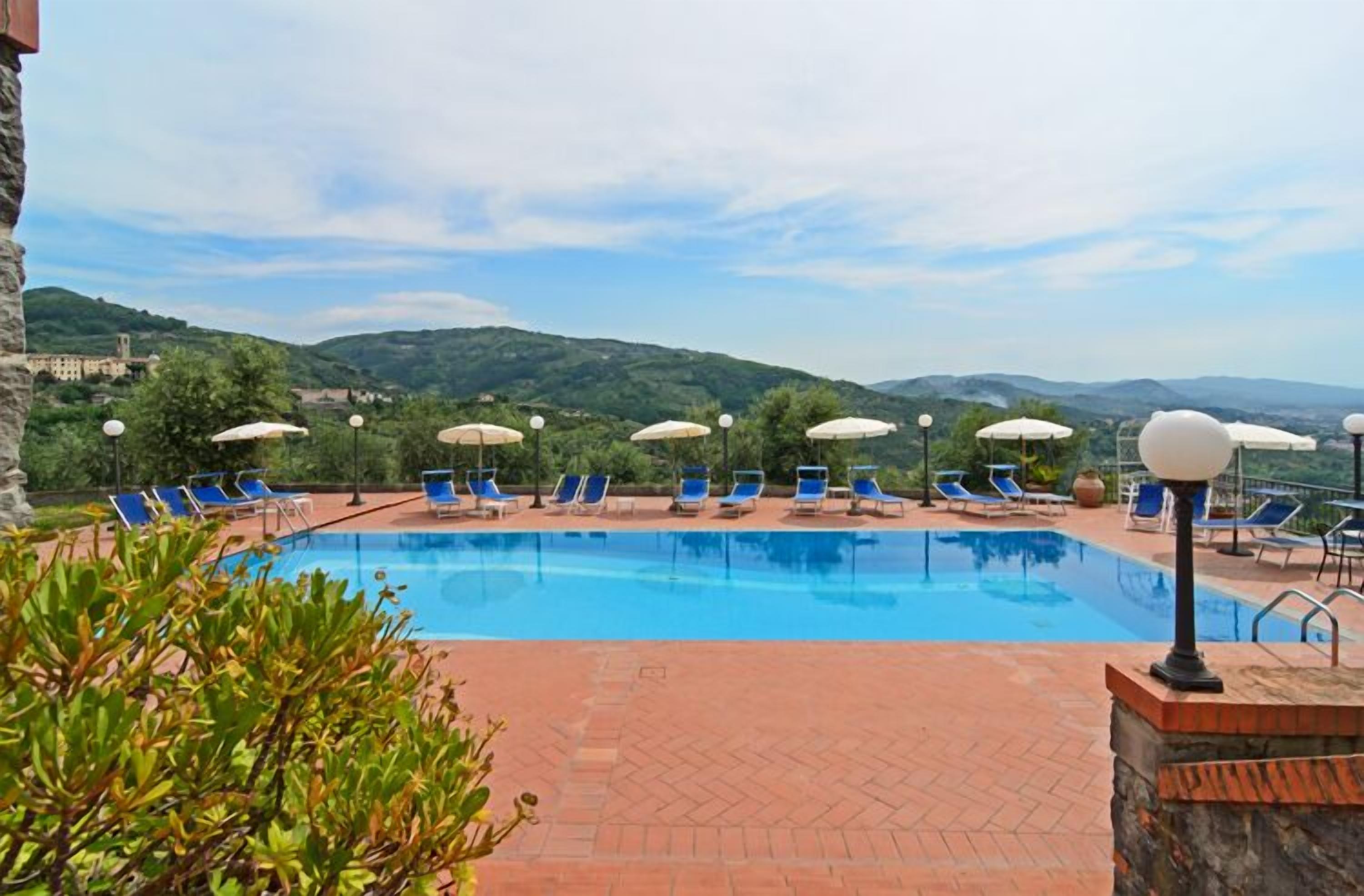 A pool area with blue lounge chairs and umbrellas, surrounded by greenery and hills in the background.