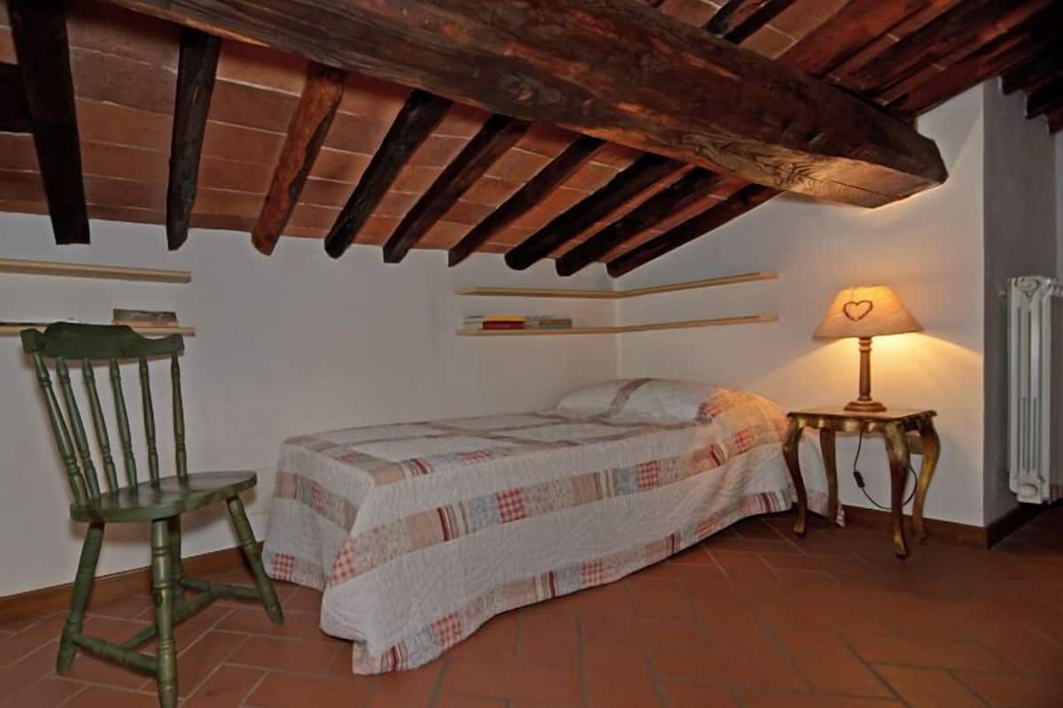 Une petite chambre à coucher accueillante, avec un plafond en bois, un lit simple, une chaise et une table de chevet avec une lampe.