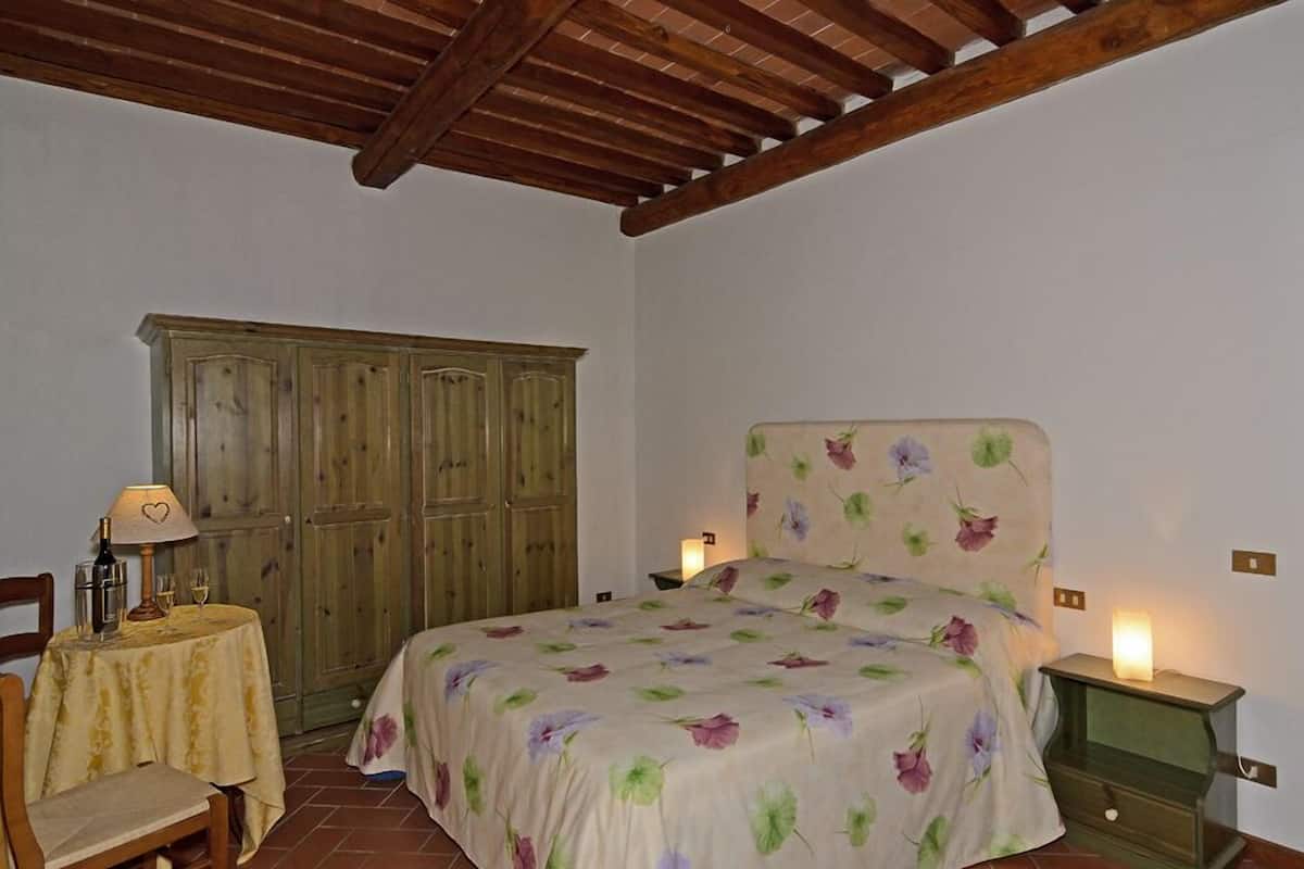 Une chambre avec un plafond en bois, un lit recouvert d’une literie à motifs floraux, une armoire en bois, une petite table avec une lampe et une table de chevet avec une lampe.