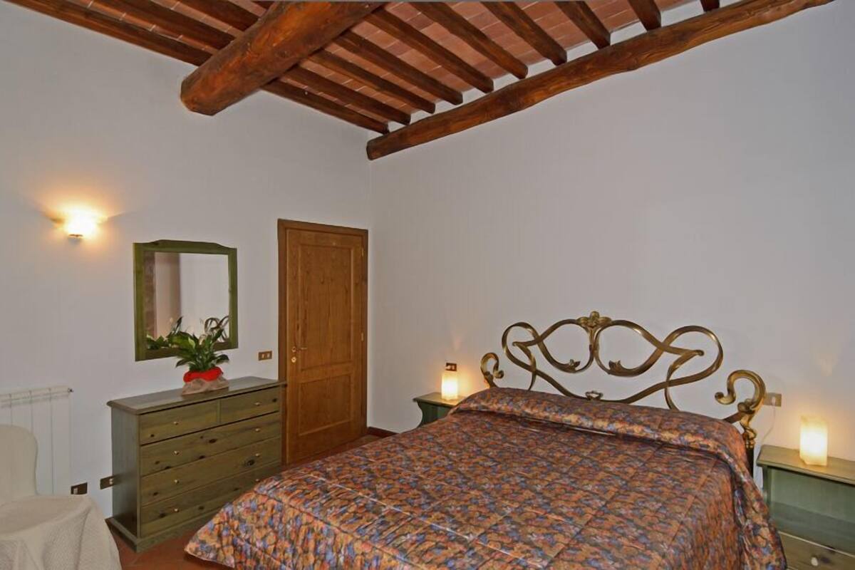 Une chambre avec un plafond en bois, un lit recouvert d’une couvre-lit à motifs, une commode, un miroir et deux lampes de chevet.