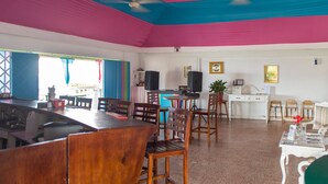 A dining area with wooden chairs and tables, a bar counter, ceiling fans, and a white desk with a computer and speakers. - Hipstrip Beach Studio (Montego Bay)