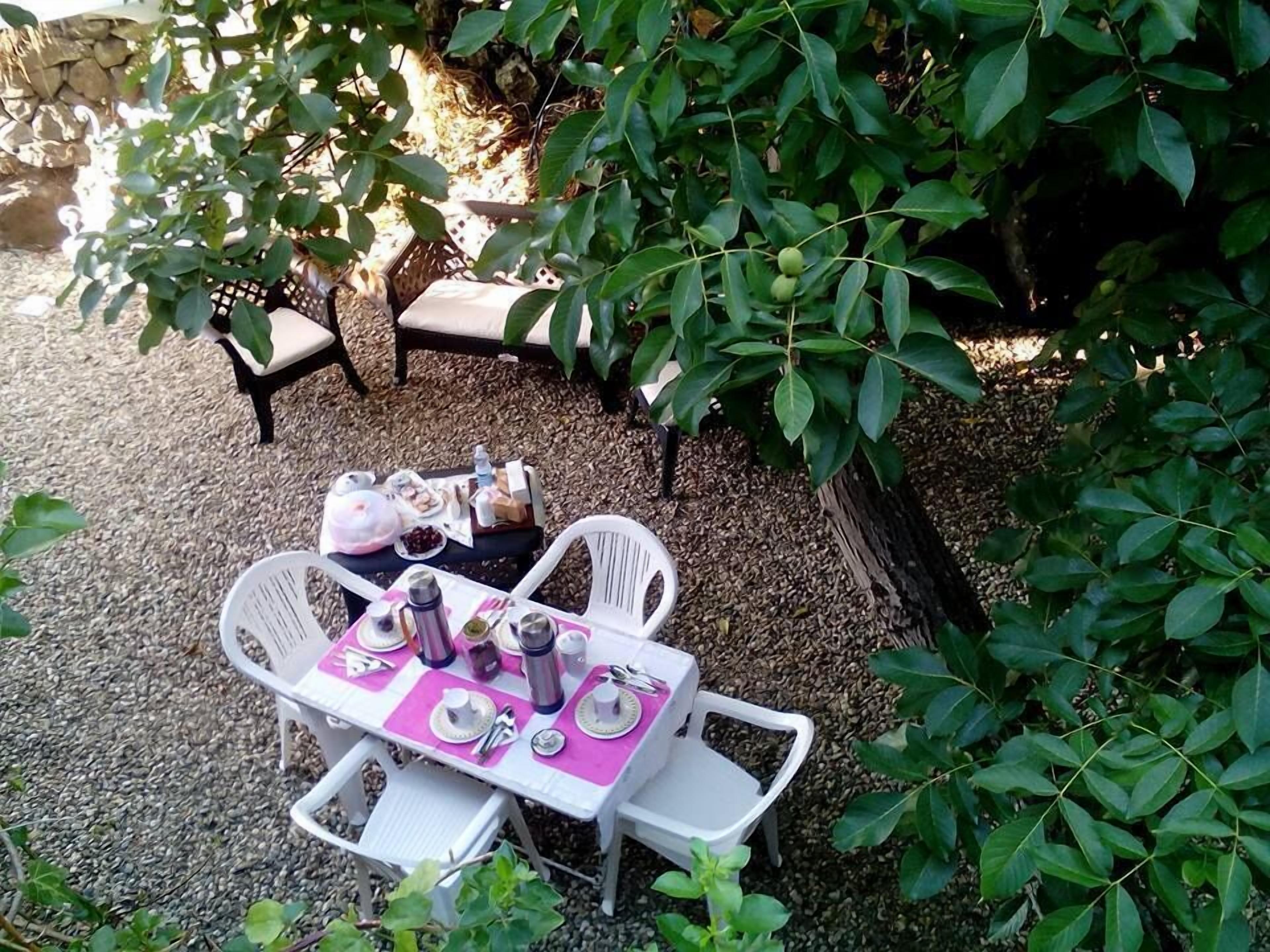 An outdoor dining area with a table set for four, surrounded by white chairs, under a tree.