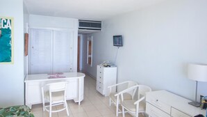 A small, clean room with a white desk, two chairs, a bed, a dresser, and a TV mounted on the wall. - Irie Beach Studio (Montego Bay)