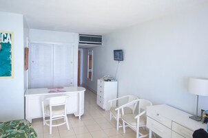A small, clean room with a white desk, two chairs, a bed, a dresser, and a TV mounted on the wall.