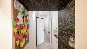 Un pasillo con un tablón de anuncios forrado con notas de colores, una puerta blanca y una pared con escritura tipo pizarra.