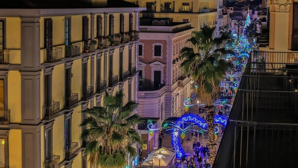 Una vista nocturna de una calle urbana bordeada de edificios, decorada con luces festivas y palmeras.