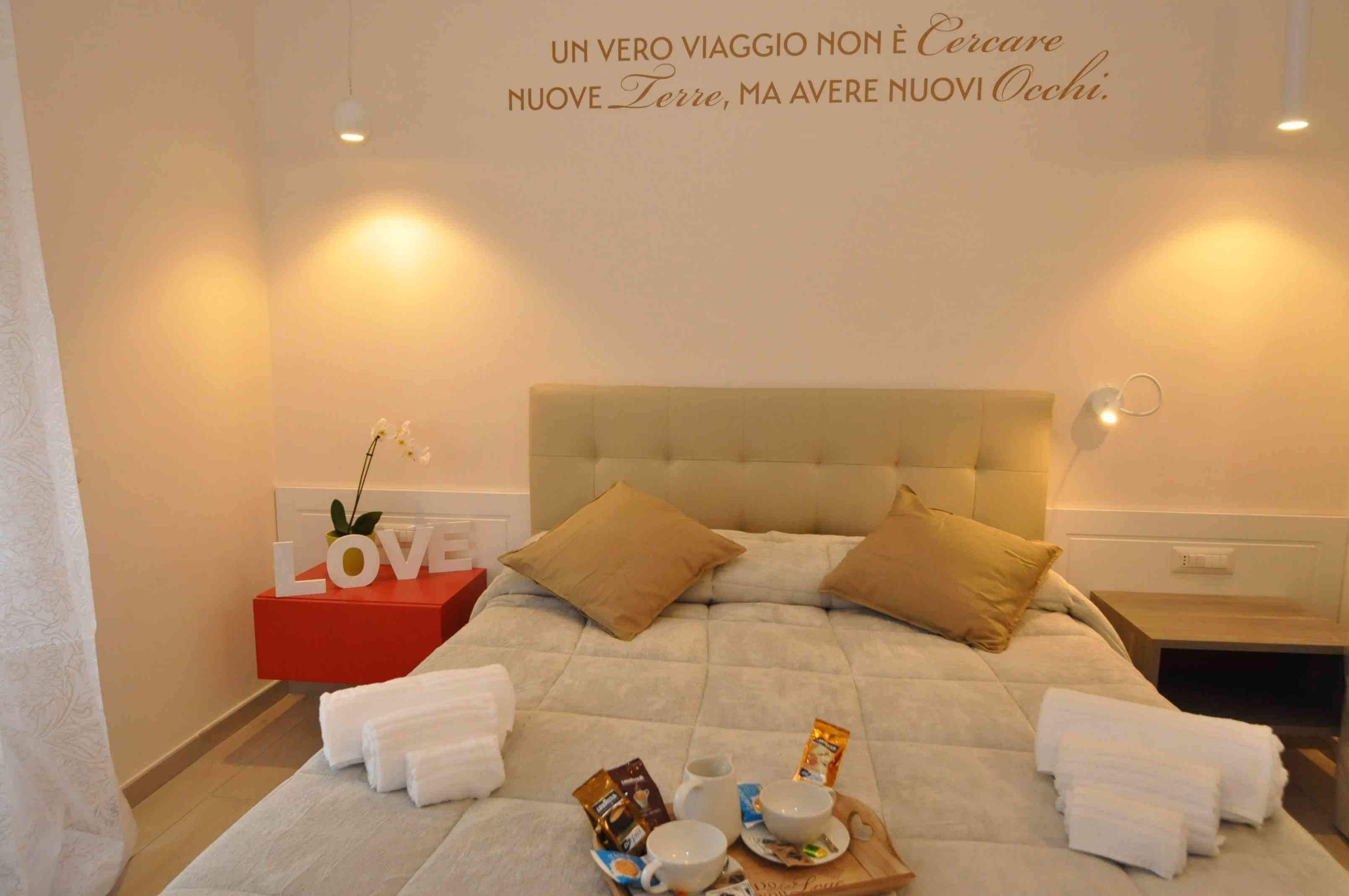 Una cama bien hecha con almohadas, una mesita de noche con lámpara y una pared con una frase motivacional.