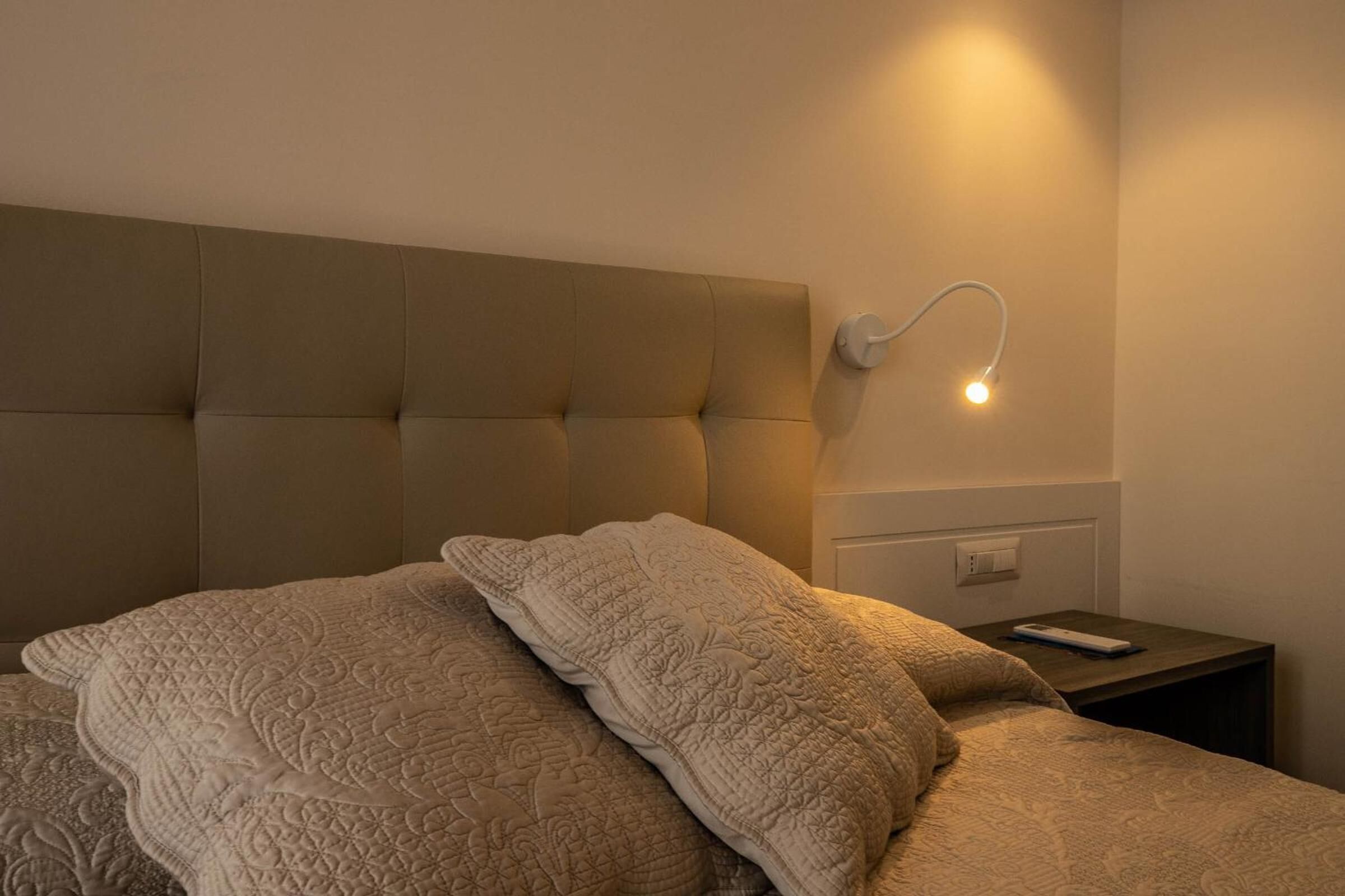 Une chambre à coucher avec une tête de lit capitonnée, une table de chevet dotée d’une télécommande et un luminaire fixé au mur.