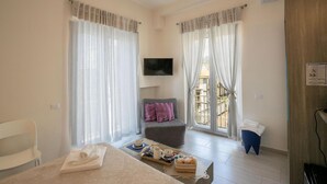 Habitación de hotel con cama, una silla, un pequeño sofá con cojines, televisor montado en la pared y un balcón con cortinas.