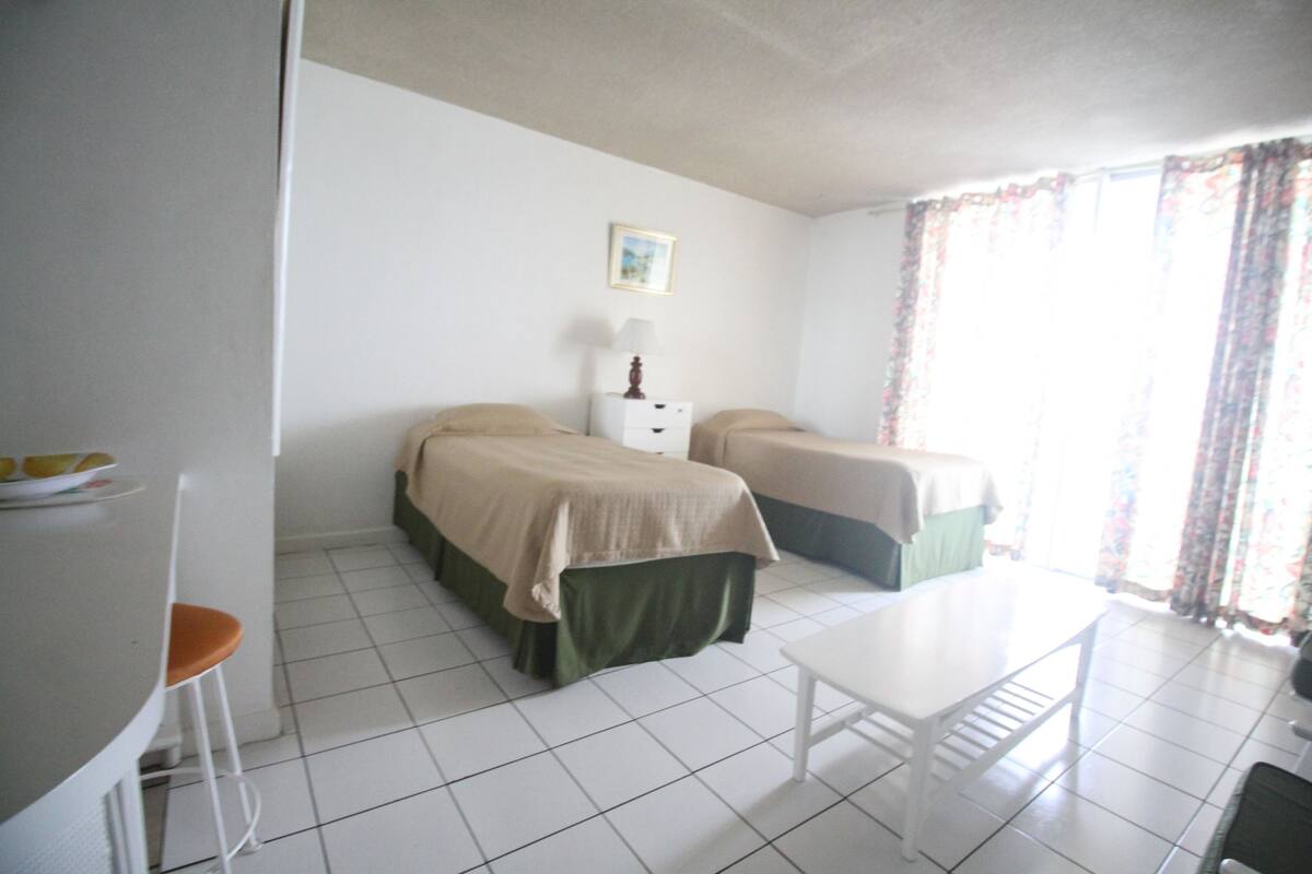 A room with two beds, a small table, a chair, and a window with curtains.
