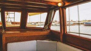 A boat's interior with wooden paneling, cushioned seating, and windows overlooking a marina with boats. - Veliero Venezia (Venice)