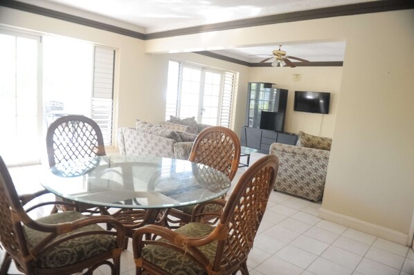 A dining area with a glass-top table surrounded by wicker chairs, a sofa set, and a flat-screen TV mounted on the wall.