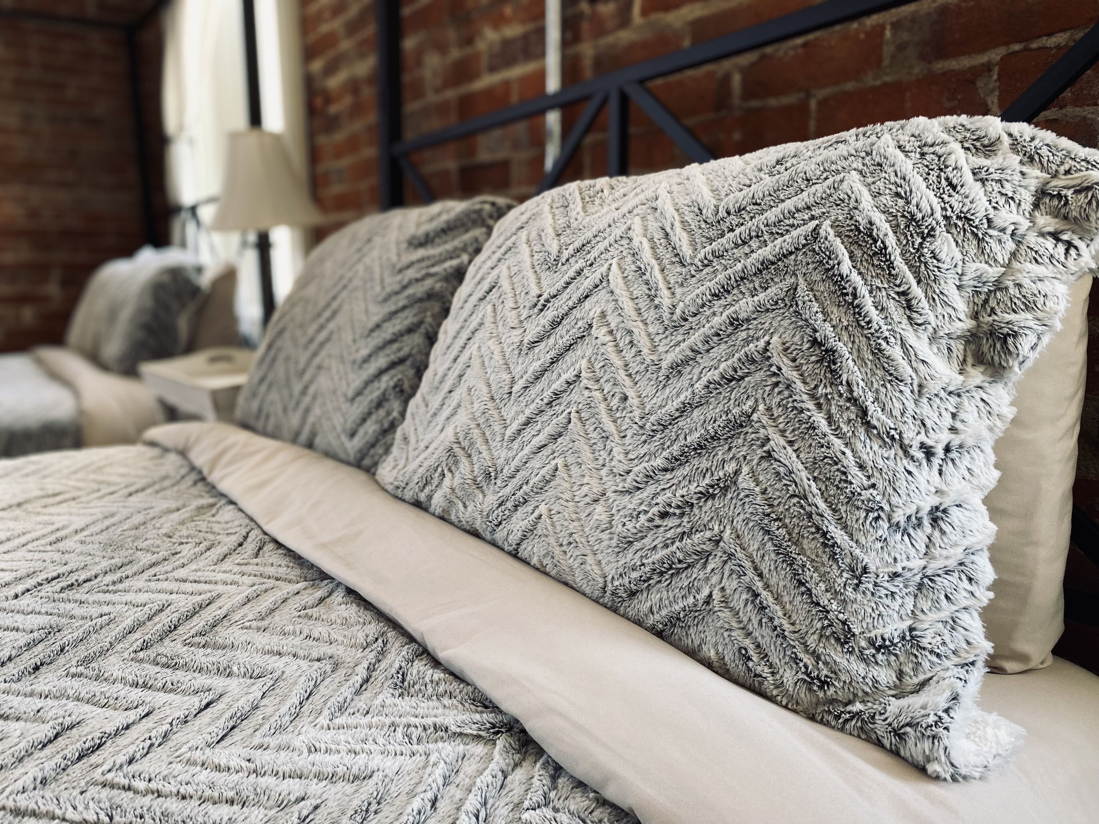 A bed with a textured blanket and pillows, a lamp, and a brick wall in the background.