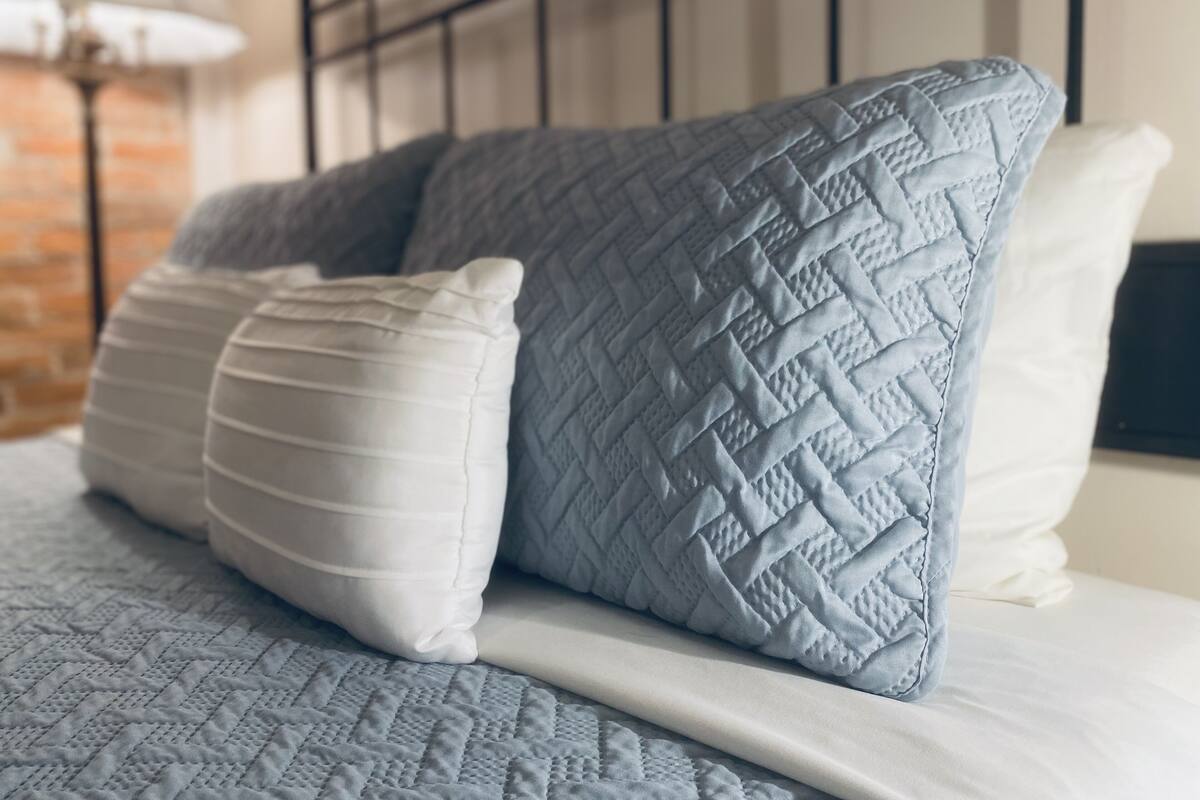 Una cama con un edredón azul acolchado y dos almohadas, una blanca y otra azul, en una habitación con pared de ladrillo y una lámpara al fondo.