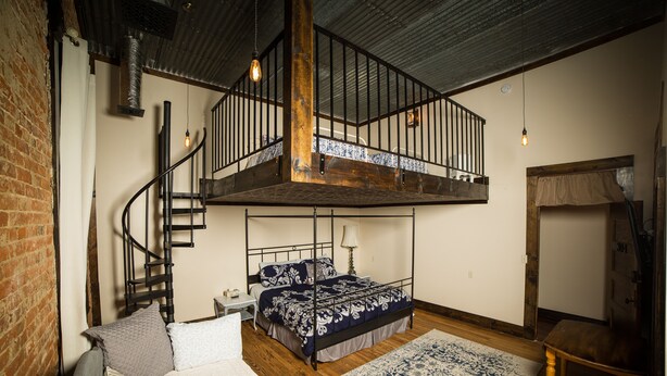 Een slaapkamer met een hoogslaper, een houten trap, een klein nachtkastje en een kamerbreed tapijt met een patroon.