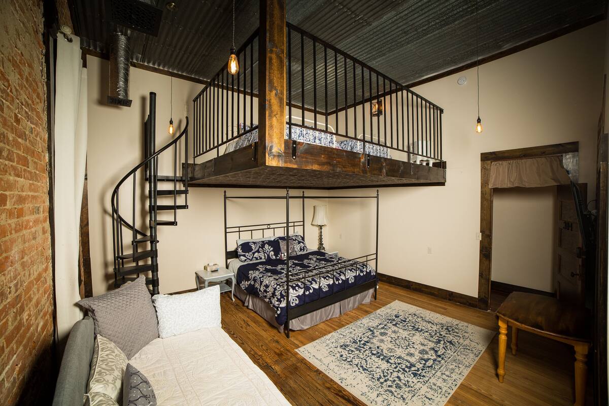 Un dormitorio con cama tipo altillo, escalera de madera, mesita de noche pequeña y alfombra estampada.