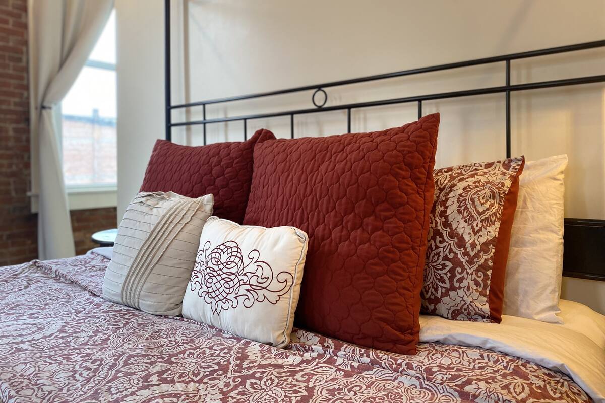 Una cama con colcha estampada, varios almohadones y cabecero de metal.